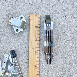 May include: A set of hardware for a cabinet door, including a silver-colored door handle, a silver-colored latch, a black knob, and a silver-colored screw. The hardware is laid out on a gray concrete surface with a wooden ruler for scale.