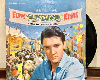 May include: A vintage vinyl record album cover featuring Elvis Presley in a blue shirt. The cover art depicts a carnival scene with a large banner that reads "Elvis Roustabout Elvis". The album is titled "The Original Soundtrack Album" and includes the text "From the Paramount Picture Roustabout".