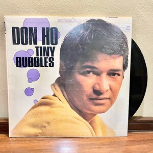 Puede incluir: Una portada de álbum de vinilo con Don Ho, un cantante hawaiano, con el título "Don Ho Tiny Bubbles" en letras grandes y en negrita. La portada del álbum es blanca con un diseño de burbujas moradas y azules. La portada del álbum es en estéreo.