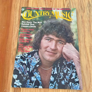 May include: A magazine cover featuring a country music singer wearing a blue floral shirt. The magazine is titled "Country Music" and features the text "Mac Davis: The Man Behind That Country Smile".