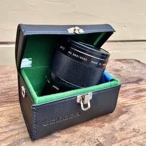 May include: A black camera lens with a green felt lining inside a black leather case. The lens has the text "MADE IN JAPAN" and "NO. 58313060" printed on it.
