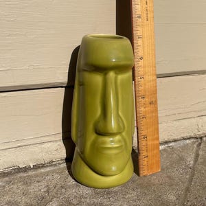 May include: A green ceramic tiki mug shaped like a face. The mug is approximately 15 cm tall.