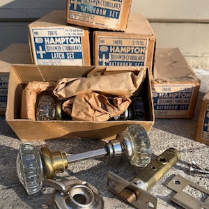 Peut inclure: Ensemble de quincaillerie de porte tubulaire Hampton Russwin vintage. L'image présente des boîtes étiquetées "Bathroom Set" et "Latch Set", ainsi que des poignées de porte en verre, des plaques métalliques et des vis. Les boîtes sont marron avec du texte bleu et blanc.