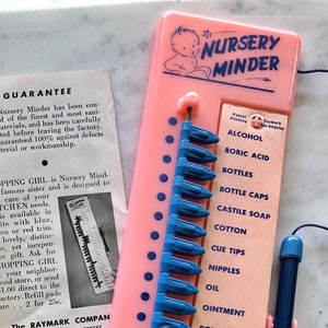 May include: A pink plastic Nursery Minder with a blue slider and a list of baby care items including alcohol, boric acid, bottles, bottle caps, castile soap, cotton, cue tips, nipples, oil, ointment, and powder.