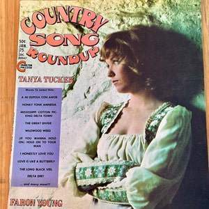 May include: A vintage record album cover featuring a young woman with long brown hair, wearing a white and green floral dress. The album title is "Country Song Roundup" with the artist's name "Tanya Tucker" in large, bold letters. The album cover is a light blue colour with white text.