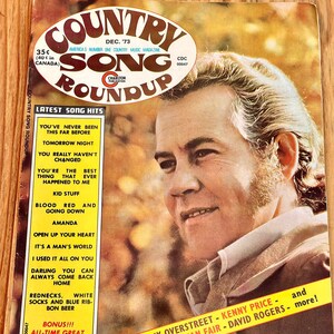 May include: A vintage Country Song Roundup magazine cover featuring a close-up portrait of a man with blonde hair and a beard. The magazine cover is yellowed with age and has the text "Country Song Roundup" in large, bold letters. The text "Dec. 73" is also visible on the cover.