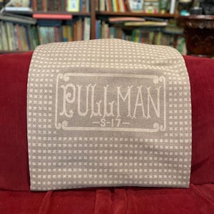 May include: A tan and white patterned pillow cover with the word "PULLMAN" in a rectangular frame. Below the word is "S-17". The pillow cover is resting on a red velvet surface.