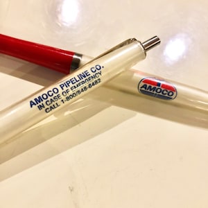 Puede incluir: Bolígrafo de plástico blanco con un logotipo de Amoco rojo y azul y el texto "Amoco Pipeline Co. In Case of Emergency Call 1-800/548-6482".