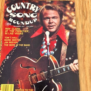 May include: A vintage Country Song Roundup magazine cover featuring Roy Clark, a country music singer, holding a guitar. The cover is red and features the magazine's title in a colourful, psychedelic font. The text on the cover reads "Country Song Roundup, December 1975, CDC 00047, Roy Clark at the Frontier in Las Vegas, Tom T. Hall, Barbi Benton, Jim Reeves, The Boys in the Band."