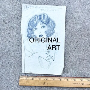 May include: A black and white pencil drawing of a woman with short, wavy hair. She is wearing a pearl necklace and holding a cigarette in her right hand. The drawing is on a piece of paper with the text "ORIGINAL ART" written in black marker.