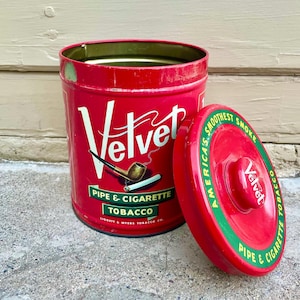 Puede incluir: Lata de metal roja con una etiqueta verde y blanca que dice "Velvet Pipe & Cigarette Tobacco" y una imagen de una pipa y un cigarrillo. La tapa es roja con un círculo verde que dice "America's Smoothest Smoke Velvet Pipe & Cigarette Tobacco".