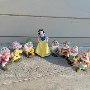 May include: A collection of ceramic figurines featuring Snow White and the Seven Dwarfs. Snow White is in a yellow dress with a blue top. The dwarfs are in various outfits, including green, yellow, and red. The figurines are arranged on a concrete surface.