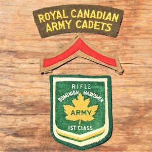 Puede incluir: Tres parches militares vintage sobre una superficie de madera. El parche superior dice "ROYAL CANADIAN ARMY CADETS" en letras amarillas sobre un fondo verde oscuro. Debajo hay un galón rojo y beige. El parche inferior es verde con una hoja de arce amarilla y texto.