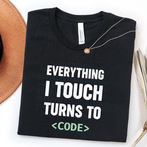 Può includere: Una t-shirt nera a maniche corte con il testo bianco "EVERYTHING I TOUCH TURNS TO" seguito dalla parola "<CODE>" in verde chiaro. La maglietta presenta un sottile motivo di cifre binarie sullo sfondo e un'etichetta bianca sul colletto.