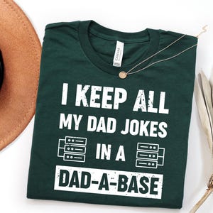 Puede incluir: Una camiseta de manga corta de color verde oscuro con texto blanco desgastado que dice I KEEP ALL MY DAD JOKES IN A DAD-A-BASE, acompañada de dos iconos gráficos que representan racks de servidores a ambos lados de la palabra IN.