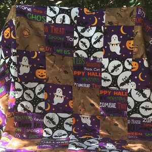 Könnte beinhalten: Ein Halloween-Patchwork-Quilt mit braunen, lila und schwarzen Quadraten. Das Design zeigt Geister, Fledermäuse, Kürbisse und Text wie "Happy Halloween" und "Spooky".