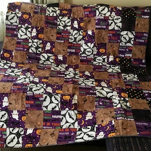 May include: A Halloween-themed patchwork quilt. The design features squares with ghosts, bats, pumpkins, and Halloween phrases. The quilt's colours are purple, brown, black, and white.