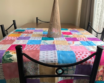 Op de afbeelding: Een patchwork tafelkleed met een verscheidenheid aan kleurrijke stoffen, waaronder roze, paars, blauw en groen. Het tafelkleed ligt op een houten tafel met twee zwarte metalen stoelen. Een decoratieve geweven kegel staat in het midden van de tafel.