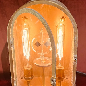 May include: A vintage-style brass lantern with two exposed light bulbs. The lantern has a rectangular shape with a hinged door and a decorative top. The interior glows with warm light, and the lantern sits on a red surface.