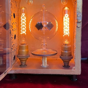 May include: A vintage-style brass and glass lamp with an open front. The lamp features decorative filigree, Edison-style light bulbs, and a central glass orb. The warm glow of the bulbs illuminates the interior, creating a cozy ambiance.