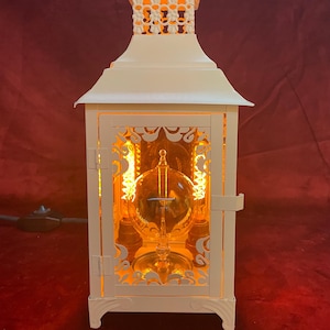 May include: A white metal lantern with a decorative top and side cutouts. The lantern has a glass globe inside, illuminated with a warm orange light. The lantern is rectangular with a handle at the top.