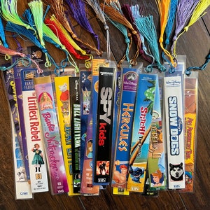 Puede incluir: Conjunto de marcapáginas hechos a mano con cintas VHS antiguas. Cada marcapáginas presenta una borla colorida y un título de película, como "La Rebelde", "Barbie y el Cascanueces" y "Hércules".