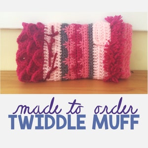 Puede incluir: Un calientamanos de crochet rosa, rojo y gris con un detalle de flecos. El calientamanos está hecho por encargo y se llama "Twiddle Muff".