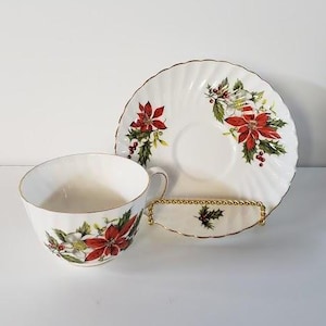 May include: A white teacup and saucer set with a gold rim and a poinsettia floral design. The teacup has a handle and the saucer is round. The set is displayed on a white surface.