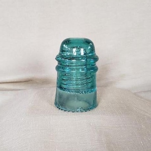 May include: A vintage, teal-colored glass insulator. The insulator has a rounded top and a ribbed design, with a flared base. The glass is transparent, allowing light to pass through. It is a classic piece of antique electrical hardware.
