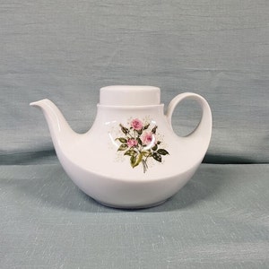 May include: White teapot with a floral design featuring pink roses and green leaves. The teapot has a curved spout and a lid.