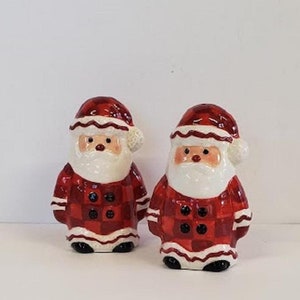 May include: A set of two ceramic Santa Claus salt and pepper shakers. The shakers are dressed in red and white plaid with white trim and black buttons.