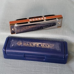 May include: A Hohner Blues Harp harmonica with a navy blue case. The harmonica is silver with a brown and gold top. The case has the words "Blues Harp" printed on it.