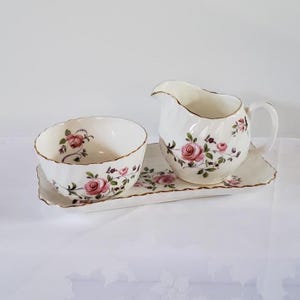 May include: A white porcelain creamer and sugar bowl set with a floral rose pattern. The set includes a creamer with a handle, a sugar bowl, and a rectangular tray. The edges are trimmed with gold.
