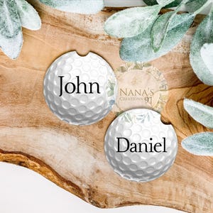 May include: Two white golf ball-shaped coasters with black text. The coaster on the left says "John" and the coaster on the right says "Daniel".
