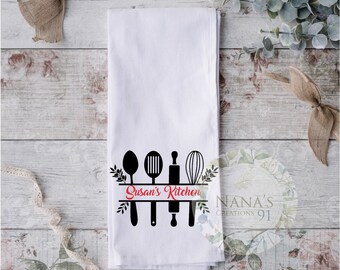 May include: White kitchen towel with a black silhouette of kitchen utensils, including a spoon, spatula, rolling pin, and whisk. The text "Susan's Kitchen" is printed in red on a black banner.