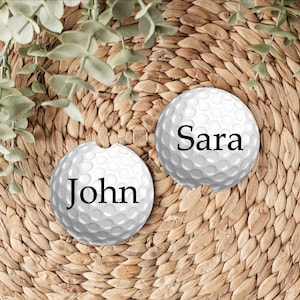 May include: Two round white car cup holder coasters with a dimpled golf ball texture. One coaster features the name John in black serif font, and the other features the name Sara in black serif font. Each coaster has a small notch on the edge.