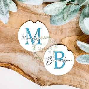 May include: Two white round coasters with blue initial letters and names. The first coaster has the letter "M" and the name "Madysen". The second coaster has the letter "B" and the name "Brandon".