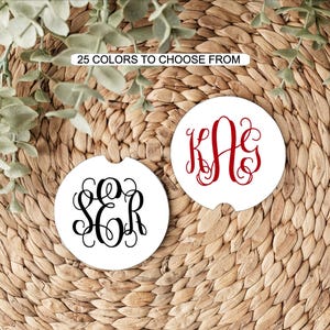 May include: Two round white car coasters featuring decorative script monograms. One coaster displays the black letters SER, while the other shows the red letters KAG. A rectangular label above the coasters states 25 COLORS TO CHOOSE FROM.