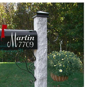 May include: A black mailbox with a white vinyl decal that says "Martin 7709". The mailbox is mounted on a gray stone post with a decorative metal bracket.