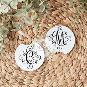 May include: Two circular white car coasters featuring black decorative script monograms of the letters C and M. Each coaster has a small semi-circular notch on the edge. The design includes intricate swirling flourishes surrounding each letter.