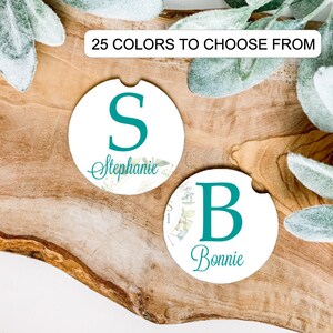 May include: Two white car coasters with teal lettering and floral accents. One coaster features the letter "S" and the name "Stephanie", the other has the letter "B" and the name "Bonnie". The text "25 COLORS TO CHOOSE FROM" is at the top.