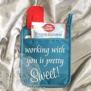 May include: Teal blue oven mitt with the text "working with you is pretty sweet!" and the text "NANA'S CREATIONS 91" in white. A red spatula and a package of Betty Crocker Sugar Cookie mix are in the pockets of the mitt.