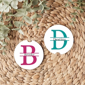 May include: Two circular white car coasters featuring personalized designs. One coaster displays a large magenta letter B with the name Brooke centered across it. The other coaster features a large teal letter D with the name Destiny centered across it.