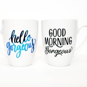 May include: Two white ceramic mugs with black lettering. One mug says "hello gorgeous" in blue and black lettering. The other mug says "good morning gorgeous" in black lettering.