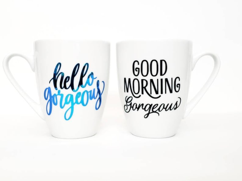 May include: Two white ceramic mugs with black lettering. One mug says "hello gorgeous" in blue and black lettering. The other mug says "good morning gorgeous" in black lettering.