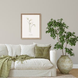 May include: A framed botanical illustration of white flowers with green stems and leaves. The print is titled "Cotta Grass" and is displayed on a white wall above a white couch with green throw pillows and a green throw blanket.