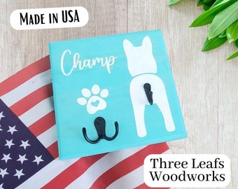 May include: A turquoise blue wooden sign with a white silhouette of a dog's rear end and a paw print with a heart. The sign has two black metal hooks and the text "Champ" and "Three Leafs Woodworks" is printed on the sign.