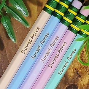 Puede incluir: Primer plano de varios lápices de colores pastel. Cada lápiz es de un color diferente, incluyendo rosa claro, azul, morado y verde. El texto "Sunset Acres" está impreso en cada lápiz en una fuente oscura. Los lápices tienen detalles verdes y amarillos.