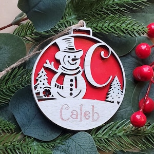 May include: A wooden Christmas ornament with a snowman design and the name "Caleb" engraved on it. The ornament is round, with a red background and a white snowman wearing a top hat and scarf. The letter "C" is also visible. The ornament is surrounded by greenery.