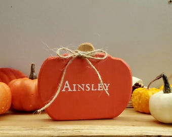 May include: A red wooden pumpkin with a twine bow and the name "Ainsley" painted in white. The pumpkin is surrounded by small gourds and pumpkins.